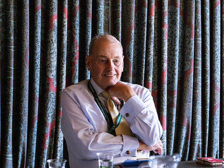 A warm portrait of an older man with thinning hair, seated at a table. He is smiling gently and looking slightly to his left. He is wearing a light blue dress shirt, a patterned gold tie, and a green lanyard. His right hand rests thoughtfully on his chin. Behind him is a backdrop of dark, pleated curtains with a red and grey pattern.