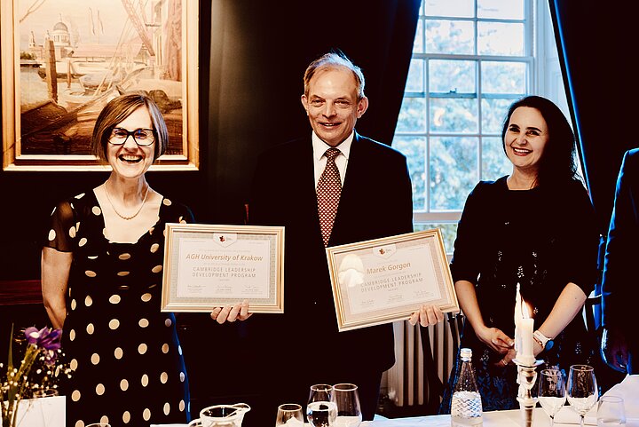 A man in a suit and two women pose for a photo at a formal dinner event. The man in the center smiles as he holds up two framed certificates for the "Cambridge Leadership Development Program". He is flanked by a smiling woman in a polka-dot dress and glasses on the left, and a smiling woman with long dark hair on the right. They are standing behind a dining table set with a white tablecloth, glasses, and a lit candle.