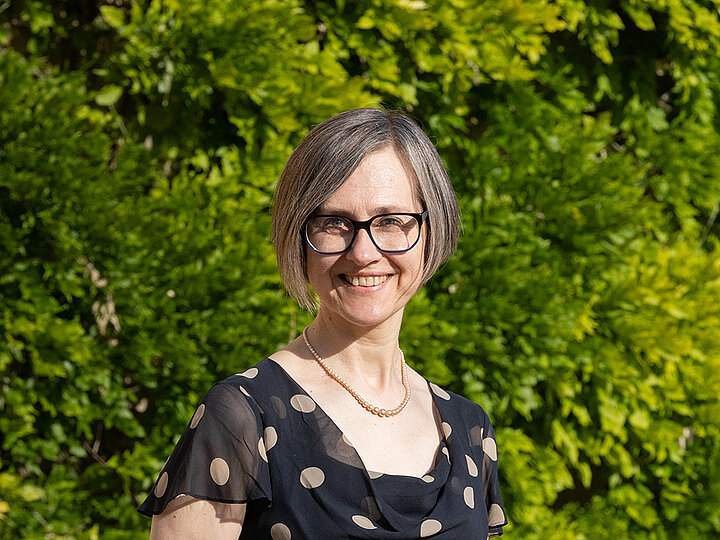  To zdjęcie przedstawia Annę Abram, która patrzy prosto w obiektyw i uśmiecha się. Ma na sobie czarną, marszczoną bluzkę w białe kropki i perłowy naszyjnik. Anna Abram ma krótkie, szare włosy i nosi okulary w czarnych oprawkach. Tło za nią stanowi bujna, zielona ściana z roślin.