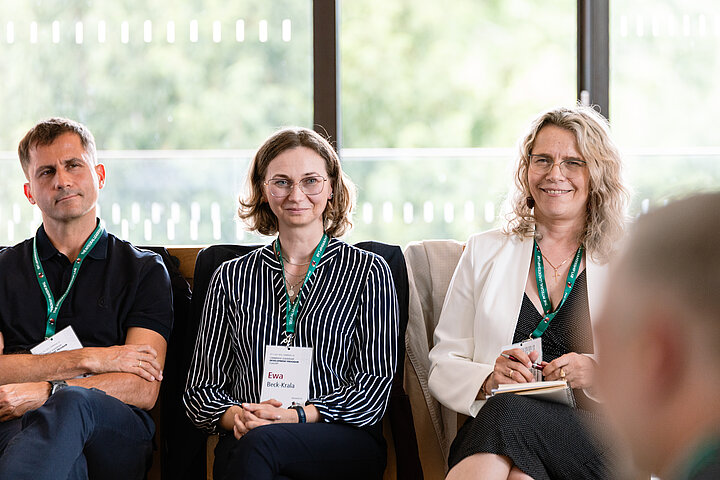 Three people seated in a row, likely at a conference or workshop, positioned in front of large windows with blurred greenery visible outside. On the left, a man with short hair wears a black polo shirt and sits with his arms crossed. In the center, a woman with shoulder-length brown hair and glasses wears a dark striped blouse and smiles towards the camera with her hands clasped. On the right, a woman with long, curly blonde hair and glasses wears a white blazer over a black top; she is smiling and looking to the right while holding a pen and notebook. All three individuals are wearing matching green lanyards around their necks.
