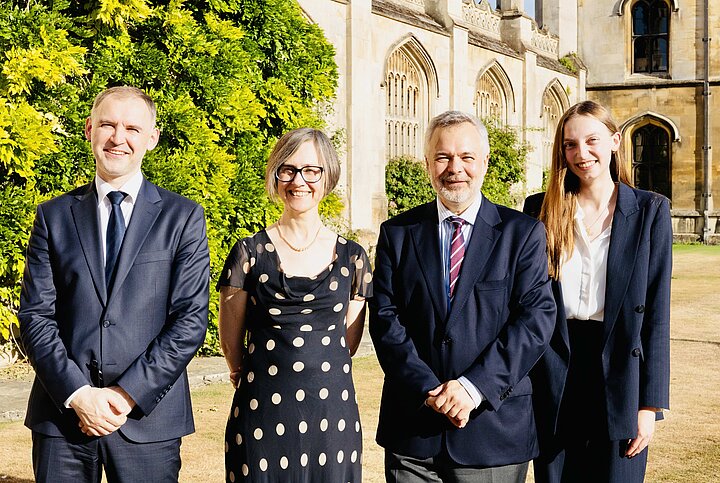 A formal group portrait of four people—two men and two women—standing side-by-side on a lawn on a sunny day. They are dressed in business attire and are smiling at the camera. In the background is a historic stone building with arched windows and a lush, leafy bush.