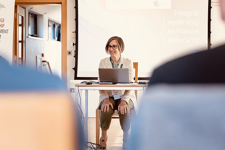 A smiling woman with short hair and glasses sits behind a desk with a laptop, appearing to lead a workshop. She leans forward with a friendly expression, engaging with the audience. Behind her, a large projection screen displays the title "Integral Leadership as Mature Leadership." The image is framed by the out-of-focus shoulders of two audience members in the foreground.