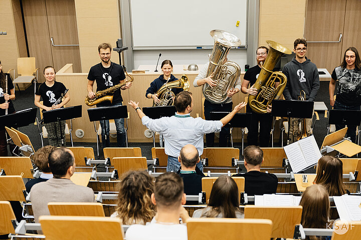 A photo taken from the back of a tiered lecture hall showing an audience watching a brass and woodwind ensemble perform at the front. A conductor stands with his back to the camera, arms wide, directing the musicians who are standing in a line holding instruments such as silver and gold tubas and saxophones.