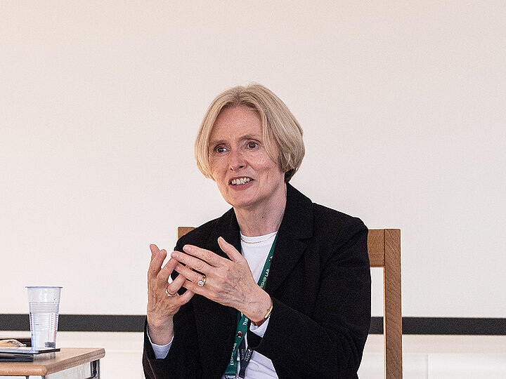 A medium shot of a woman with short, blonde hair, who appears to be speaking at an event. She is wearing a black blazer over a white top with a lanyard around her neck. She is seated and gestures with her hands as she talks, wearing an engaged and thoughtful expression. The background is a simple white wall with a thick black horizontal stripe.