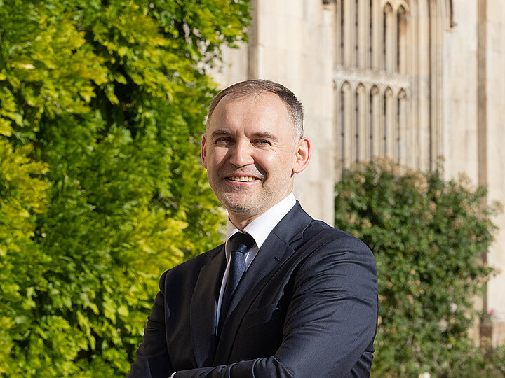 To zdjęcie przedstawia Piotra Janasa. Mężczyzna stoi, z rękami skrzyżowanymi na piersiach, uśmiecha się i patrzy prosto w obiektyw. Ma na sobie granatowy garnitur, białą koszulę i krawat w tym samym kolorze. W tle widać fragmenty zabytkowej, kamiennej fasady budynku oraz bujną, zieloną roślinność.