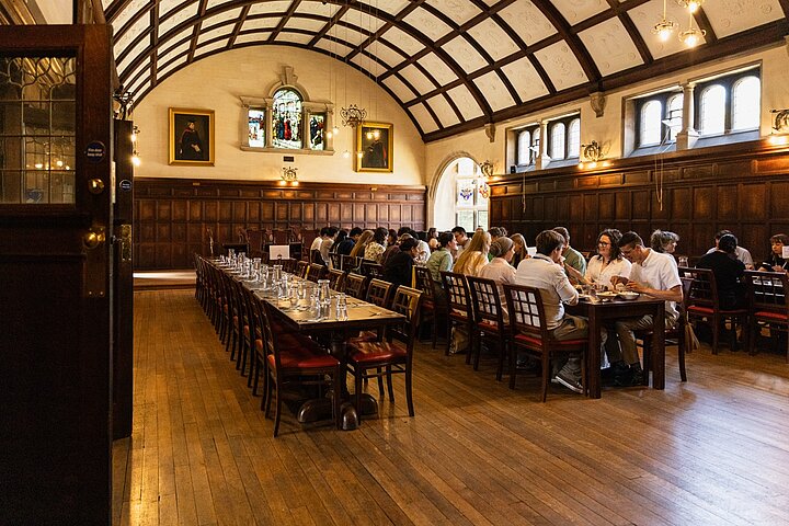 A wide view from the entrance of a grand, traditional college dining hall with a high, arched, and coffered ceiling. The walls are covered in dark wood paneling, and the floors are polished wood. A group of people are seated along long dining tables on the right side of the hall, eating and conversing. In the foreground on the left, another long table is neatly set with glasses but is currently empty. Gilt-framed portraits hang on the far wall.