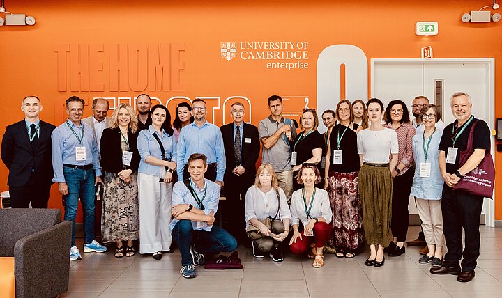 A large group of adults poses for a photo in a modern office lobby. They are arranged in standing and kneeling rows in front of a bright orange wall that features the logo for the "UNIVERSITY OF CAMBRIDGE enterprise". The participants are dressed in smart-casual attire and most are wearing conference lanyards.