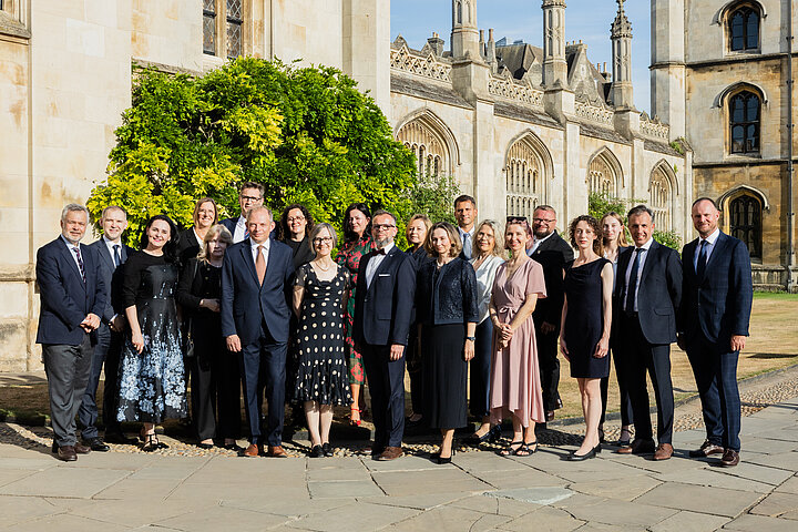 A group of approximately twenty adults posing for a formal outdoor photograph on a stone-paved courtyard. The group consists of men in dark suits and ties and women in professional dresses and business attire. They stand in front of a historic, light-colored stone building with Gothic architectural details, including arched windows and spires, with a large green bush visible behind the group on the left side.