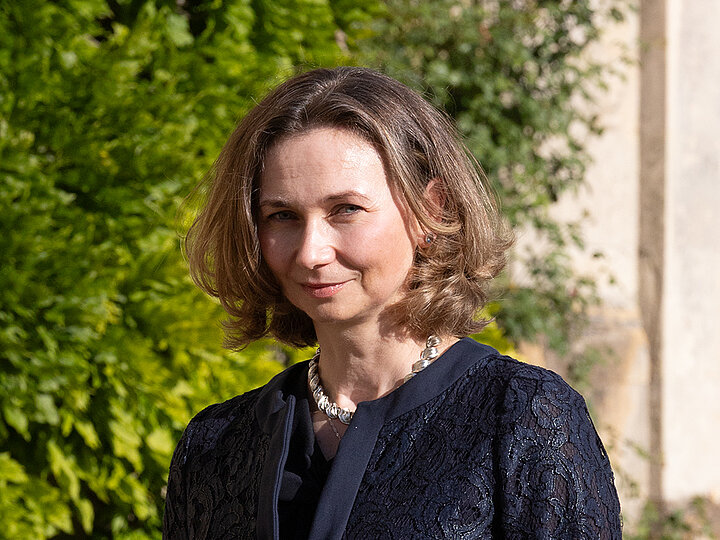 Na zdjęciu jest Ewa Beck-Krala. Kobieta ma na sobie granatową, koronkową marynarkę, a na szyi srebrny naszyjnik. Patrzy wprost w obiektyw i delikatnie się uśmiecha. Jej włosy są krótkie, a w prawym uchu widoczny jest mały, srebrny kolczyk. Tło stanowią zielone liście i fragment kamiennej, jasnej ściany.