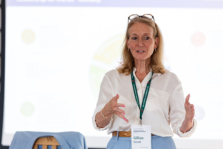 A woman with shoulder-length, light-brown hair and glasses resting on top of her head is standing and giving a presentation. She is wearing a white silky blouse, light blue trousers, and a green lanyard with a conference ID badge. She gestures with both hands as she speaks with an engaging expression. Behind her is a large white projection screen showing a faint, colorful chart.